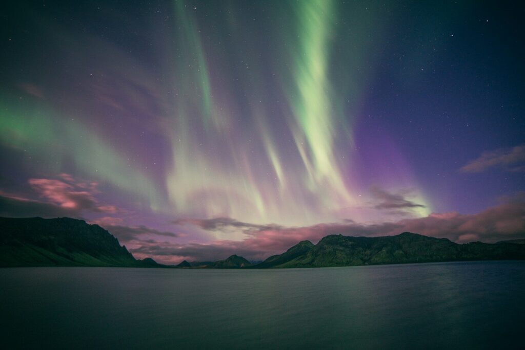 Green and purple aurora borealis above a dark lake and distant mountain range